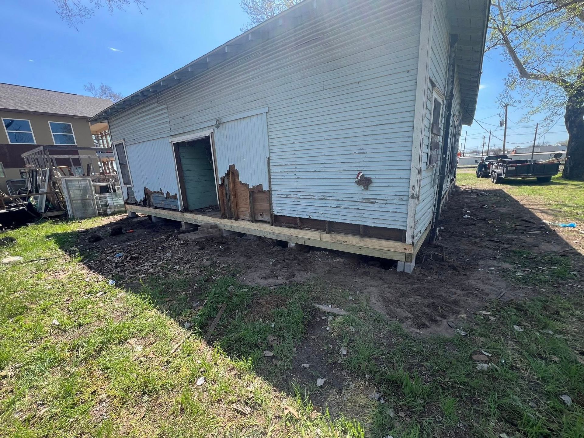Damaged white shed with leaning wall and broken siding on a grassy lot near houses