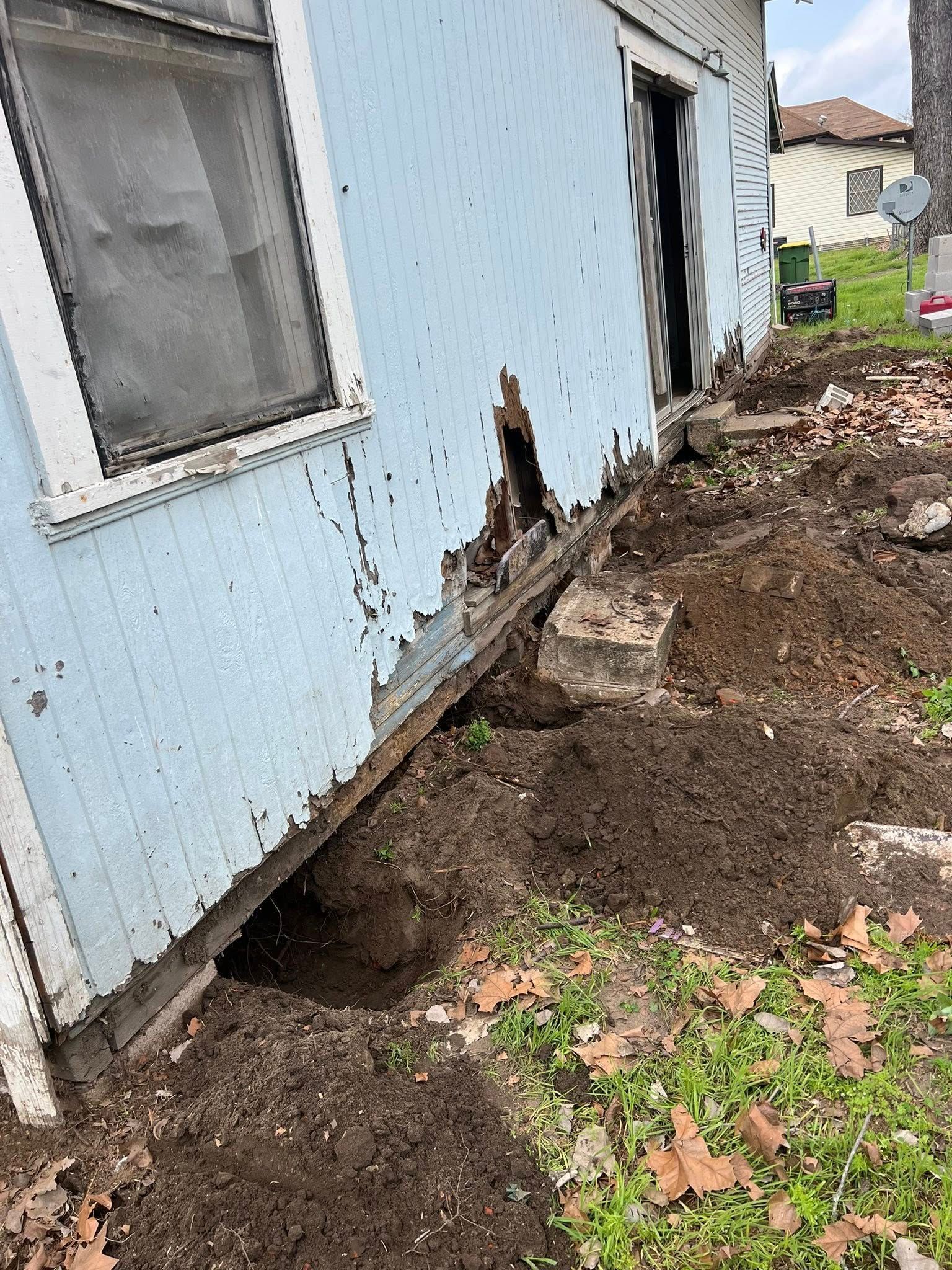 Damaged blue house siding with a large hole at the foundation beside a muddy excavation area
