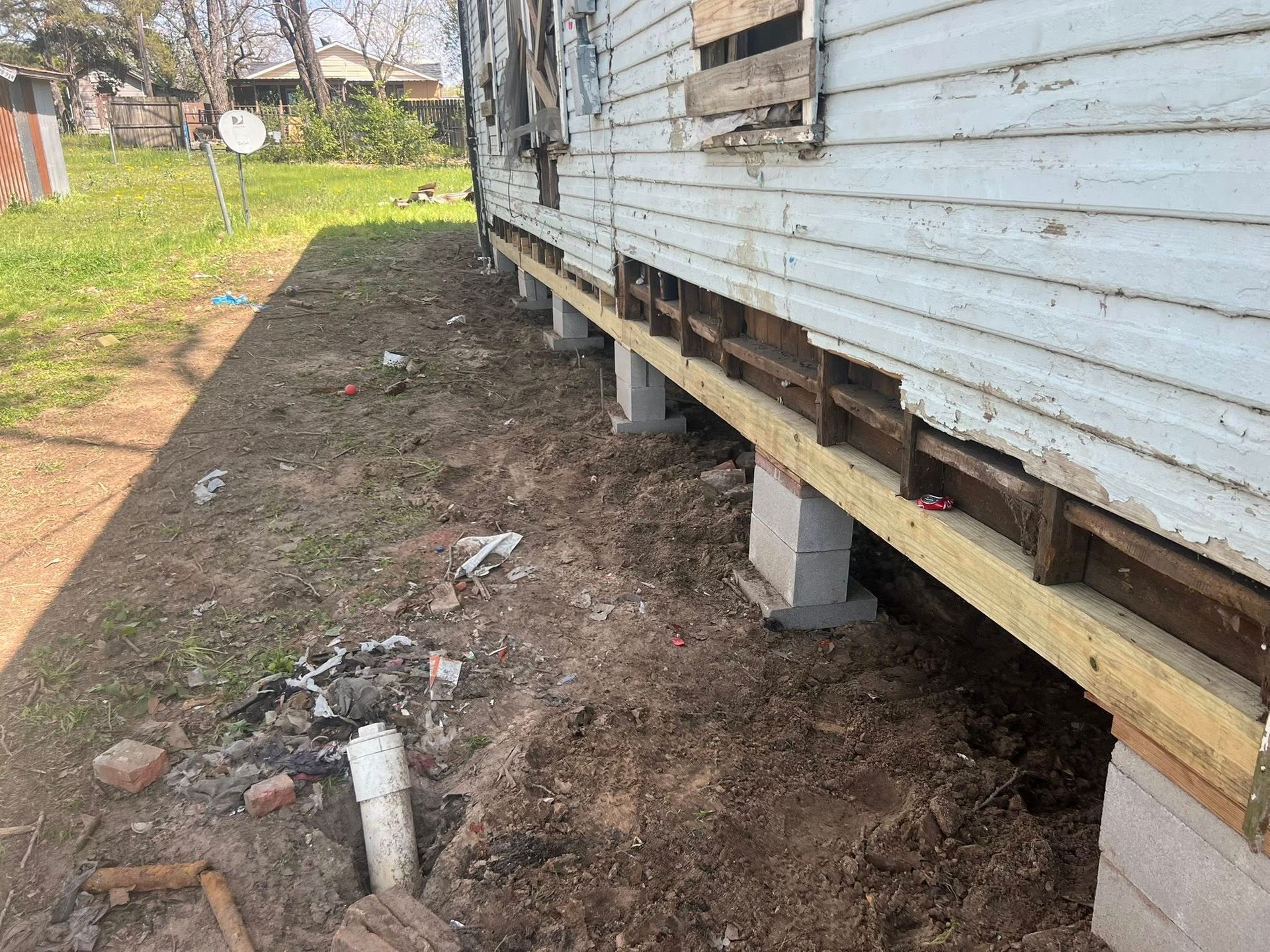 Raised white house siding with exposed foundation, showing dirt ground and support blocks along the side.