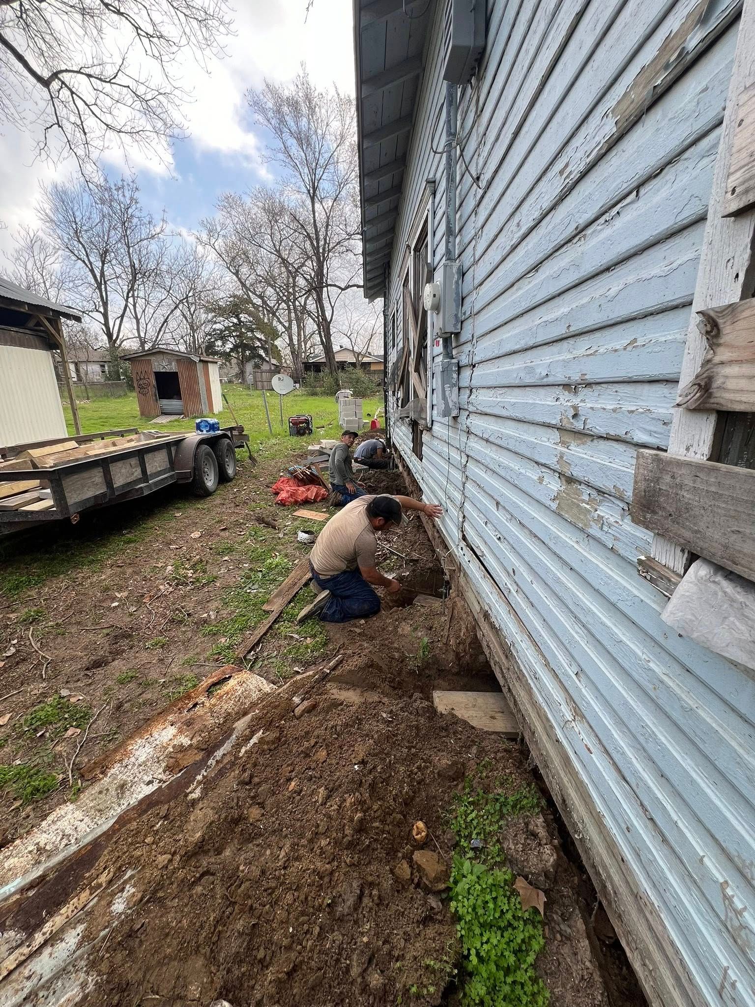 Worker digging a trench beside a blue house, with soil piled along the foundation and a trailer in the yard.