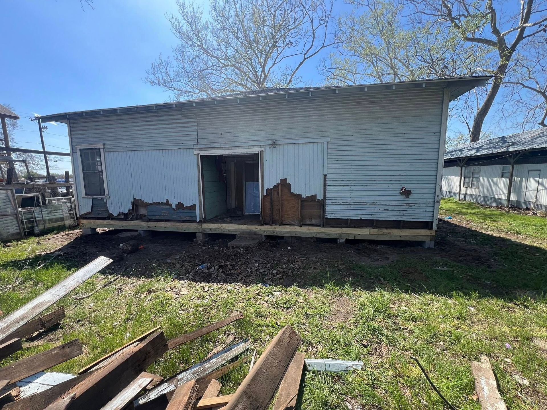 Weathered white shed with open doorway and damaged lower siding in a grassy yard, wood debris in foreground.