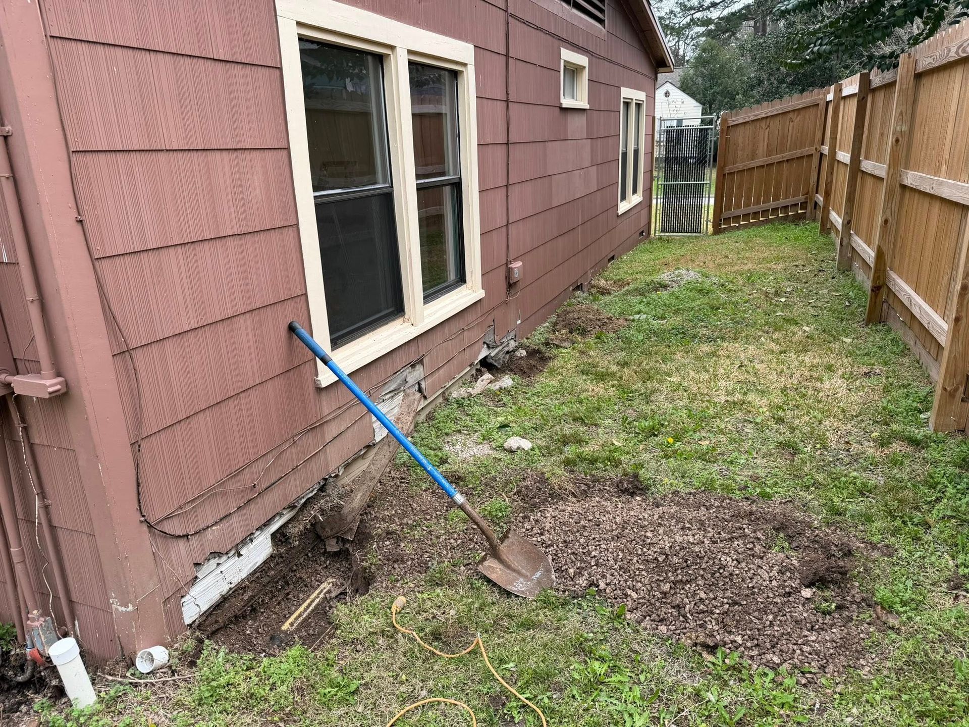 Side yard beside a pink house with a leaning blue shovel and a dirt pile near the foundation