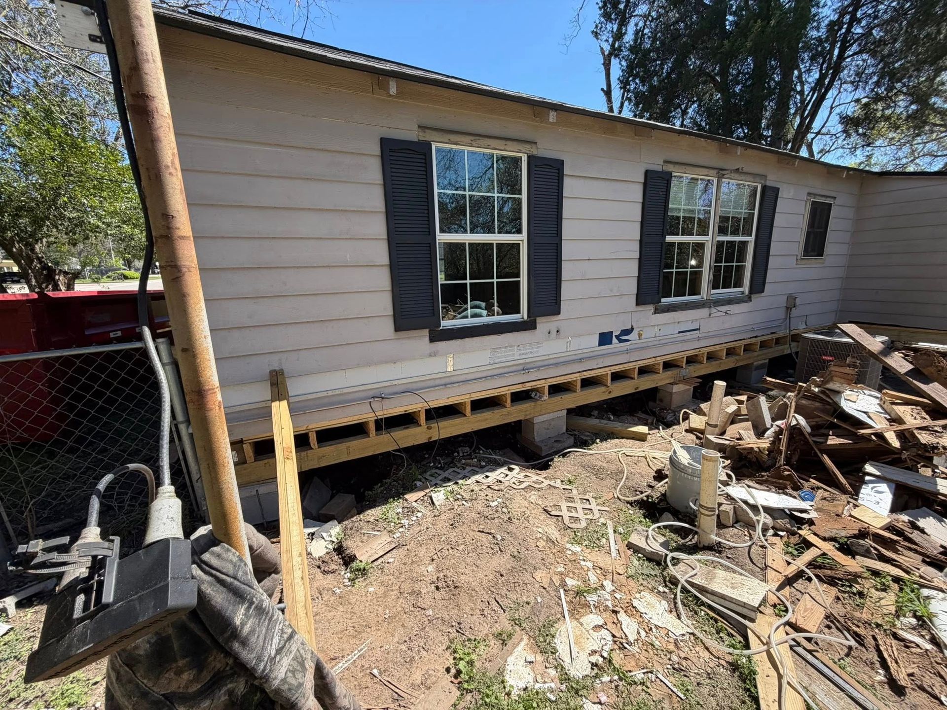 House exterior under renovation with exposed foundation, piled debris, and black shutters on white siding