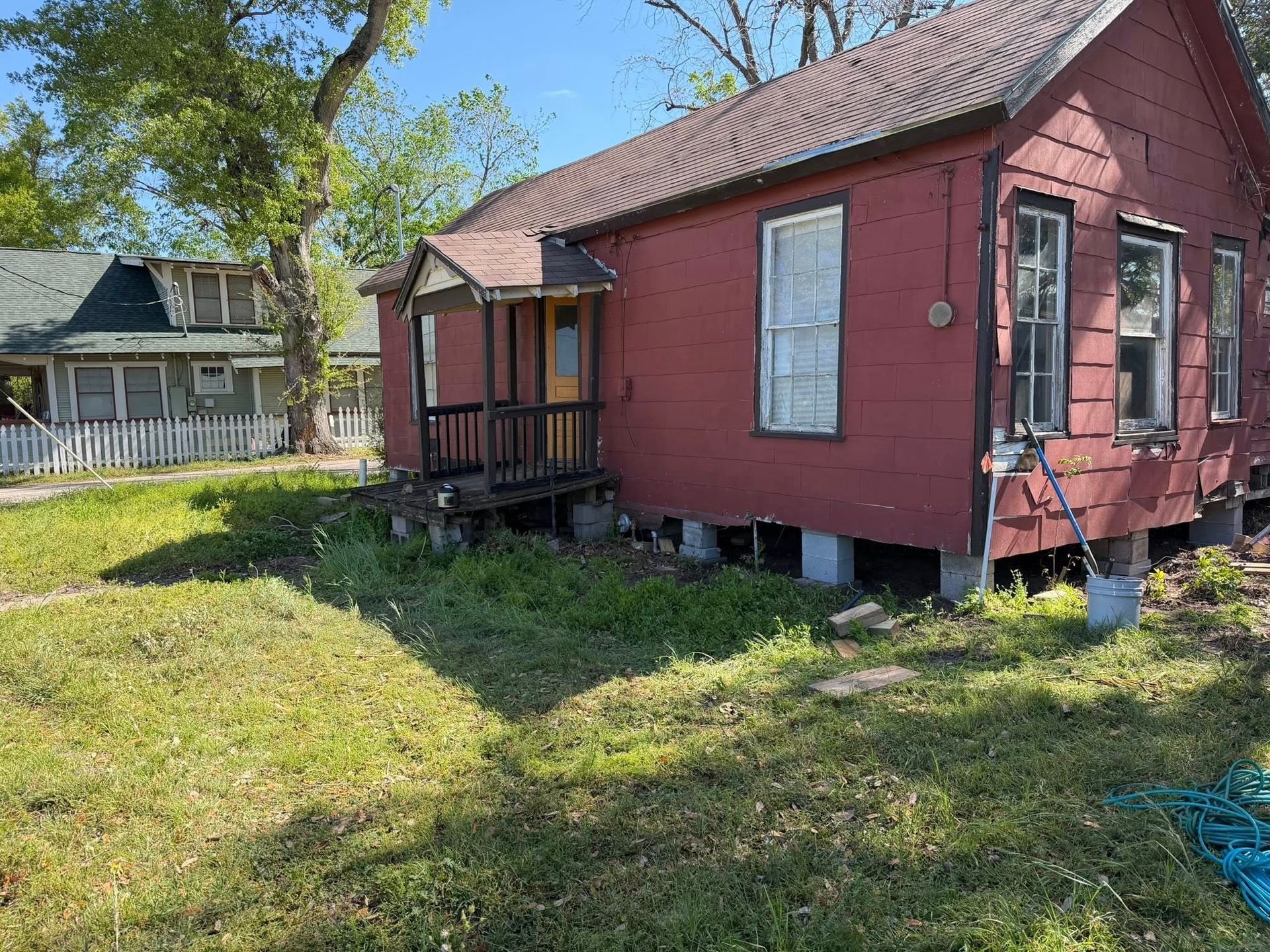 Small red shed-like house with porch in a grassy yard, with a white house in the background.