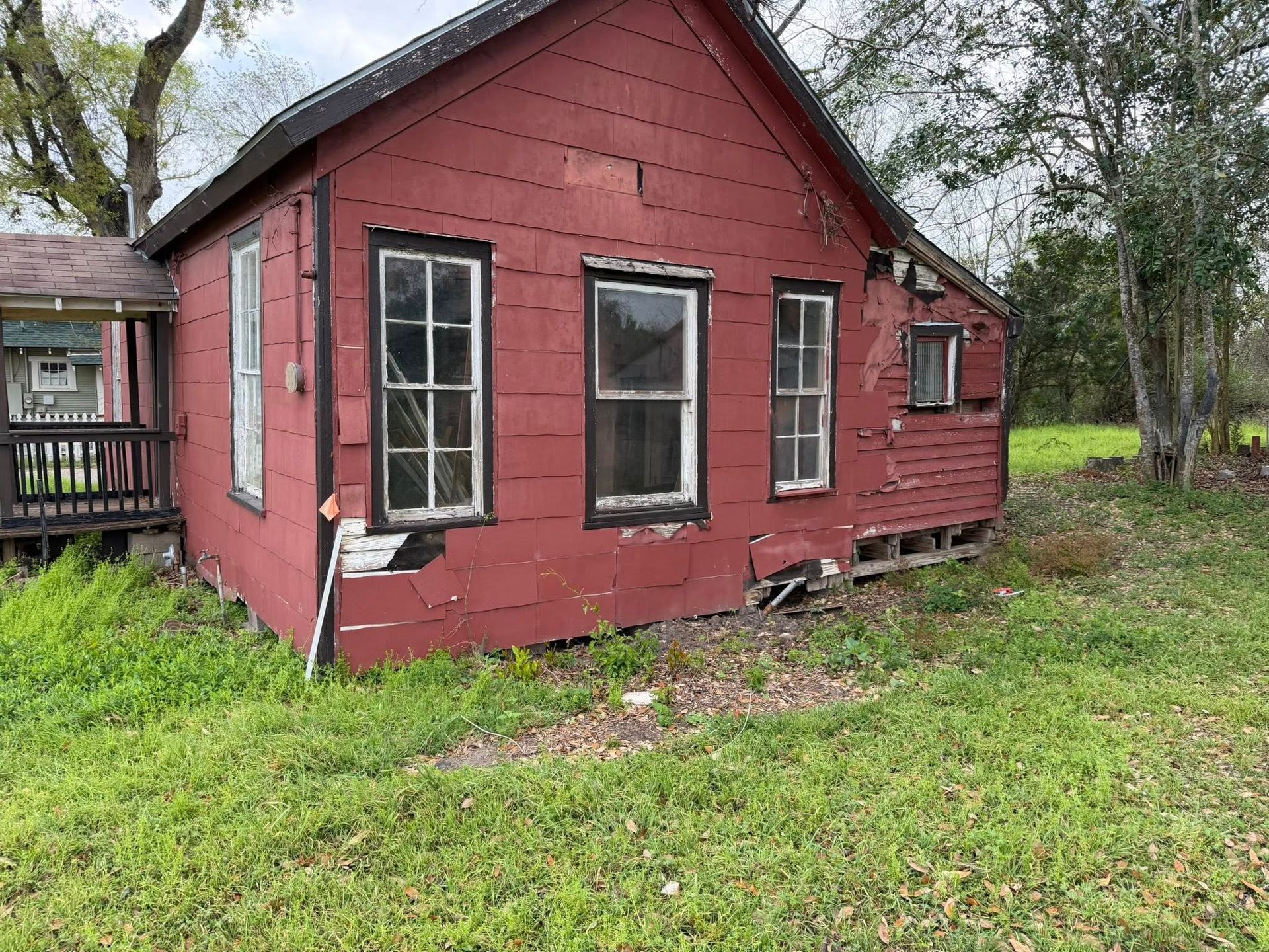 Old red house with peeling paint and broken windows, surrounded by overgrown grass and trees.