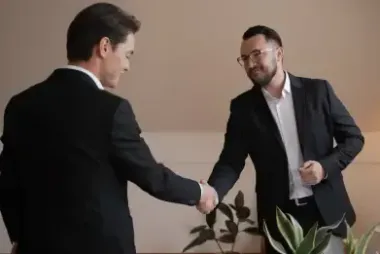 Two businessmen in dark suits shaking hands in an office setting, with plants in the foreground.