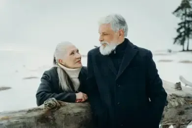Two people in winter coats stand by a log in a snowy landscape, looking at each other