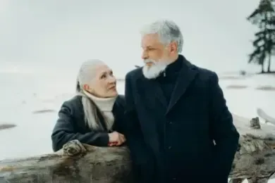 Two people in winter coats stand by a log in a snowy landscape, looking at each other