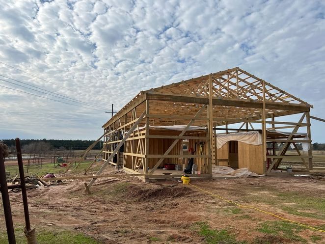 Wooden barn frame under construction on a dirt lot beneath a cloudy sky