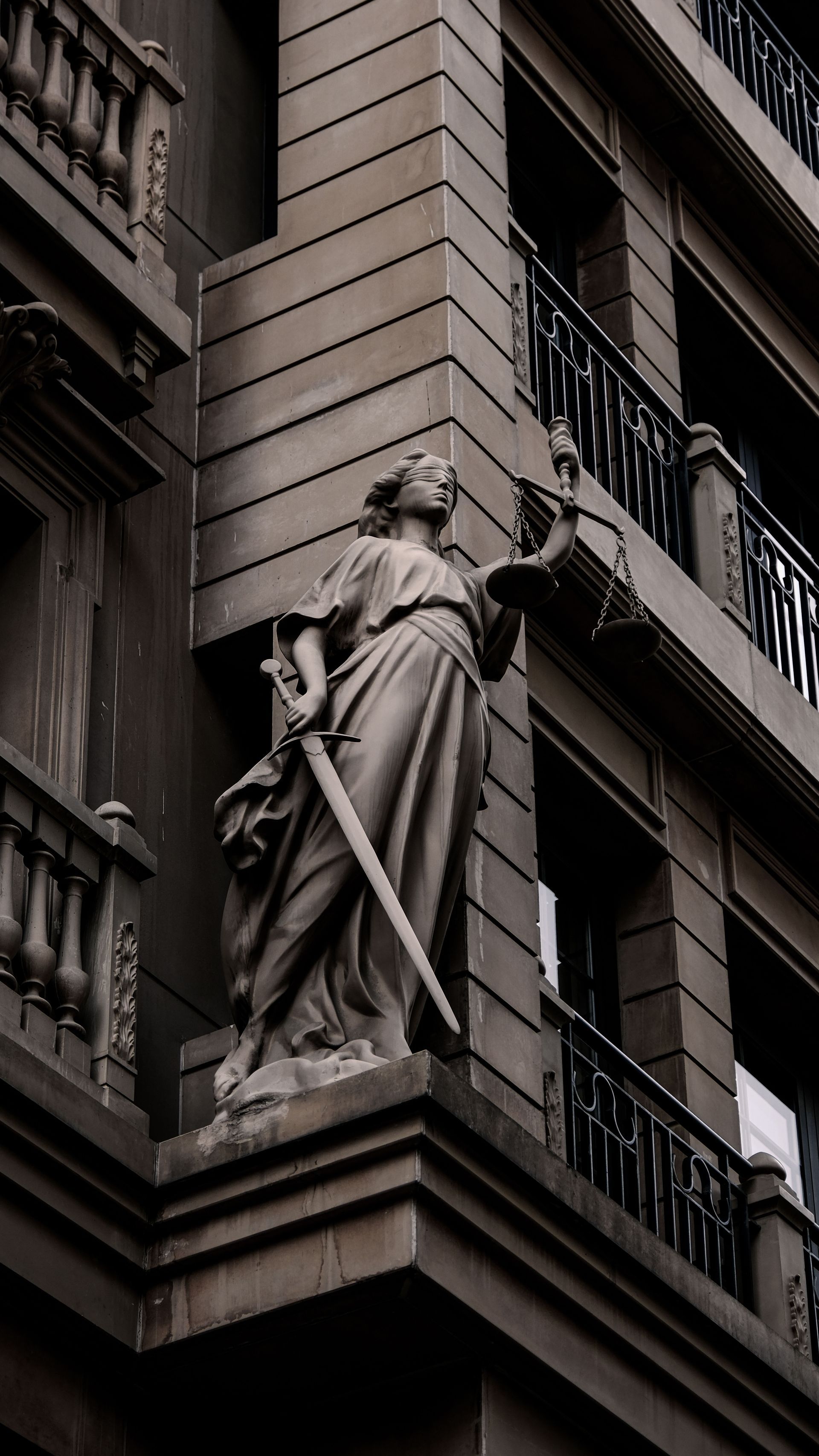 Stone statue on a building corner facade, holding a sword against dark brick architecture.