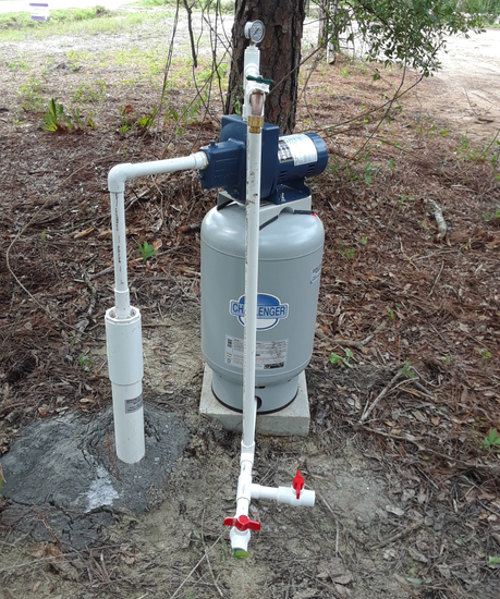 Deep Well For Fetching Water — Palatka, Fl — Moore’s Well Drilling A well pump system with a blue pump, pressure tank, and white PVC pipes outdoors.