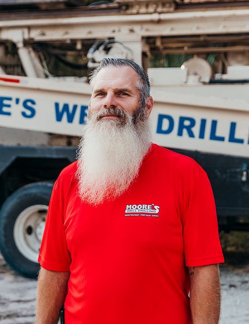Man with long white beard in red shirt stands in front of a well drilling truck.