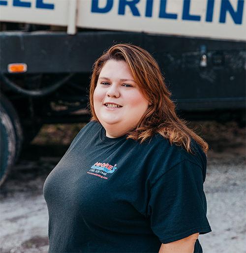Woman in a black t-shirt smiles in front of a drilling truck.