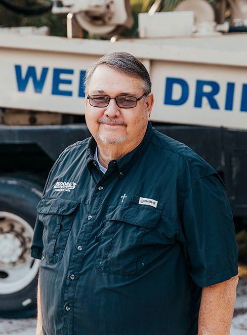Man in a dark blue shirt and glasses standing in front of a well drilling truck.