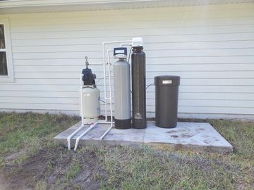 Water filtration system on a concrete pad outside a house, consisting of tanks and pipes.