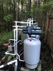 Well water system with pump, pressure tank, and pipes outdoors in a wooded area.