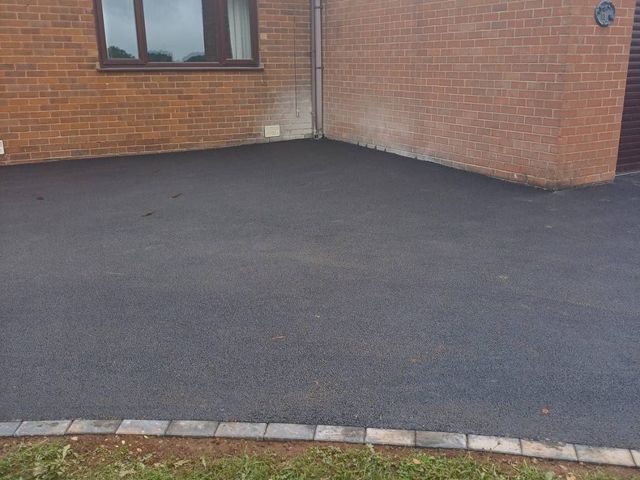 New black tarmac driveway meeting the corner of a brick house, with a block edge along the lawn