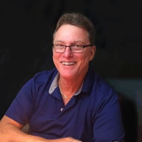 A man, Stephen Foster, with glasses smiling, wearing a blue polo shirt, sitting with a dark background.