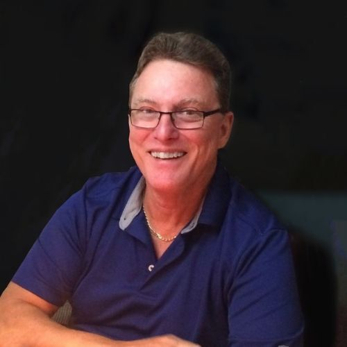 A man, Stephen Foster, with glasses smiling, wearing a blue polo shirt, sitting with a dark background.