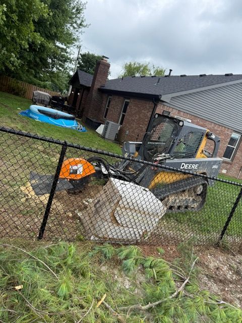 A John Deere skid steer with a stump grinder attachment works in a backyard near a house and chain-link fence.