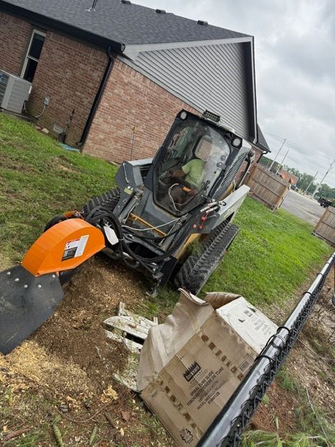 A person in a tracked skid steer with a stump grinder attachment works on a lawn next to a brick house and a fence.
