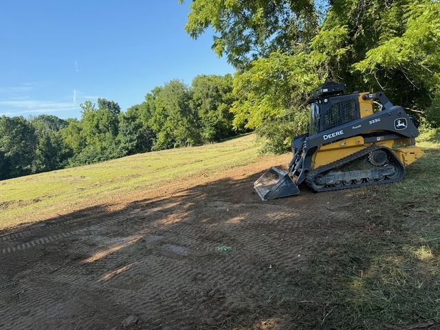 A yellow and black John Deere track loader sits on a dirt patch in a grassy field next to a tree line under a blue sky.