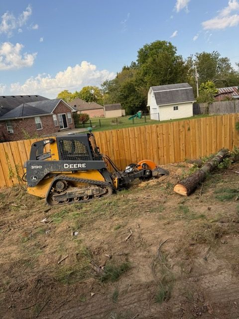 A yellow John Deere skid steer with a stump grinder attachment is working on a tree stump in a grassy backyard.