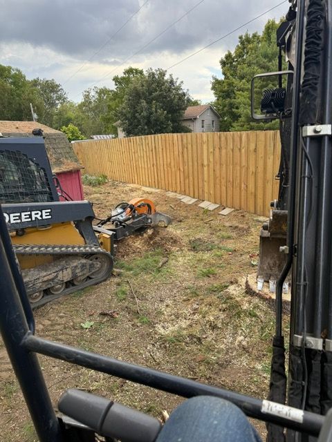 View from a construction cab showing a yellow John Deere skid steer with a stump grinder attachment working in a yard.