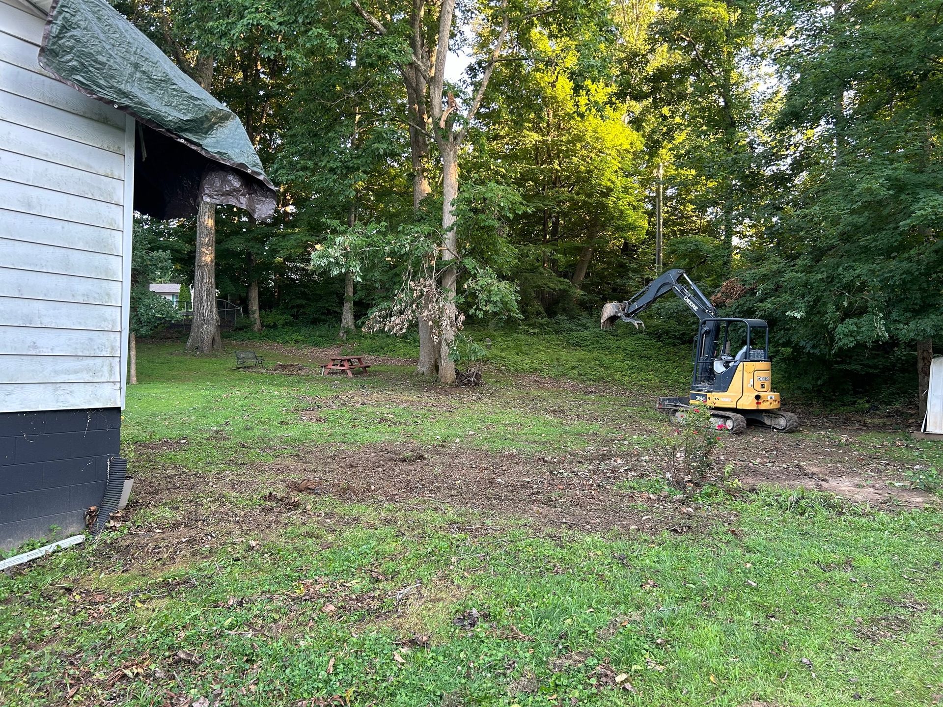A yellow mini excavator sits in a grassy, partially cleared backyard next to the corner of a white house.