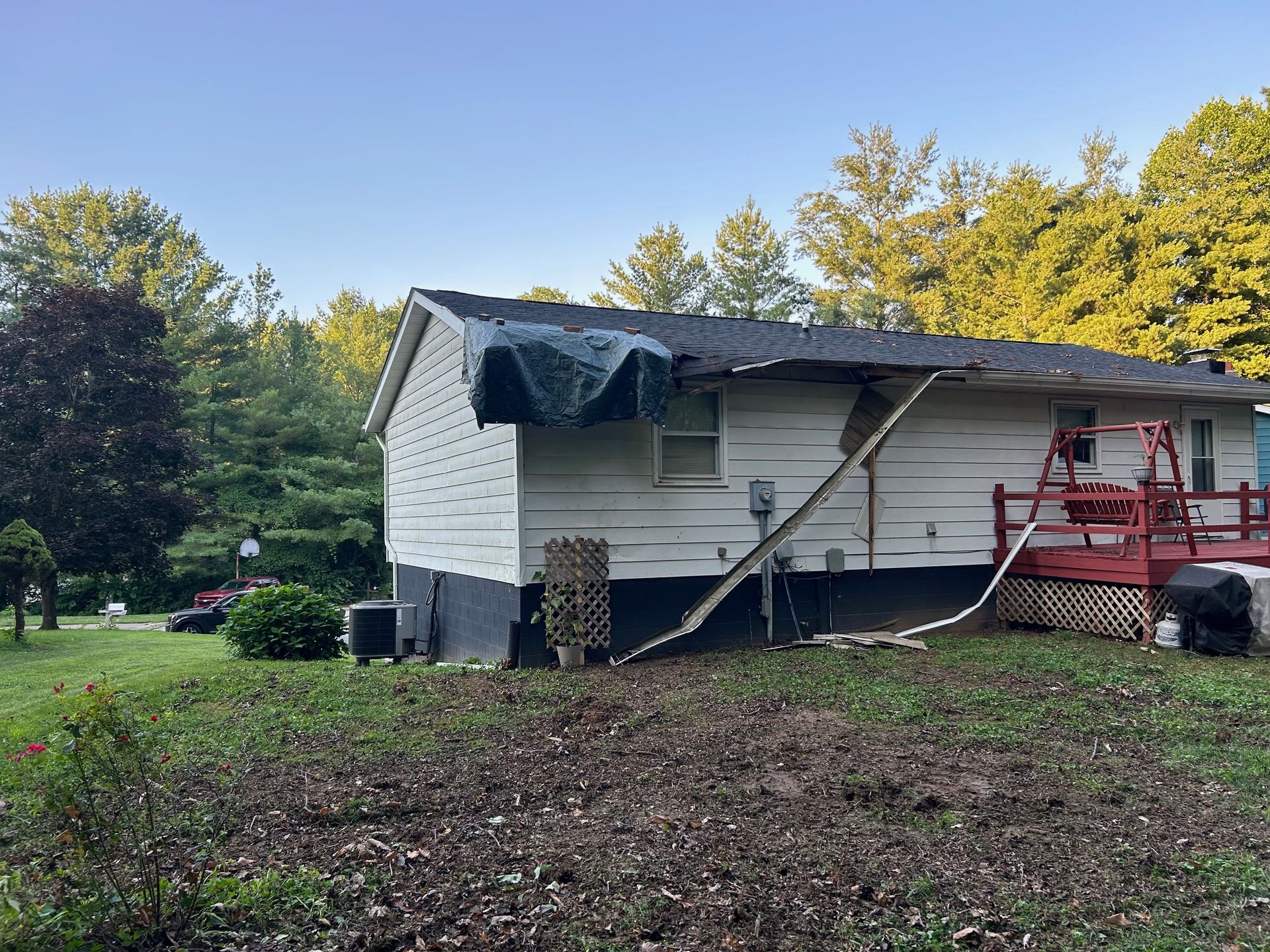 A white house with a damaged roof, featuring a blue tarp covering a section, a ladder leaning against it, and a red deck.