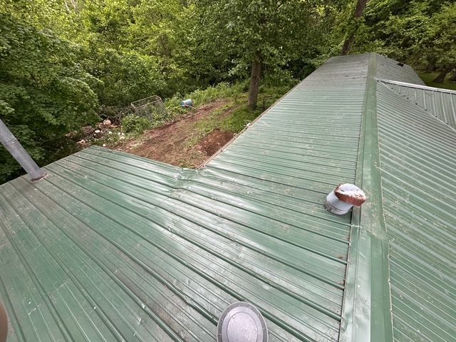 A high-angle view of a green metal roof on a building, partially surrounded by trees and a dirt yard.