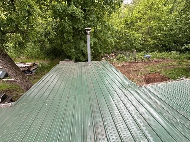 A green corrugated metal roof with a chimney pipe, viewed from above, surrounded by trees and a dirt yard.