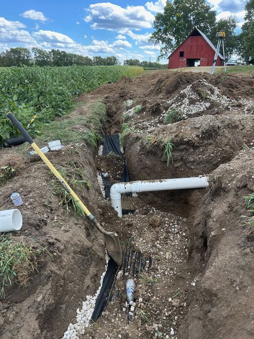 A trench dug in a farm field containing a white PVC pipe and black corrugated drainage tubing, with a shovel nearby.