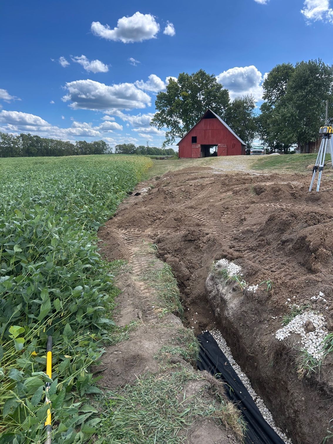 A trench with a black drainage pipe sits beside a green field, leading toward a red barn under a blue, cloudy sky.