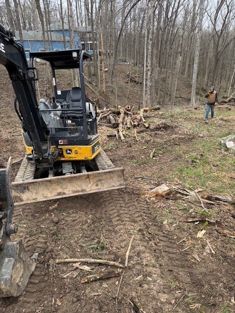A yellow John Deere mini excavator sits in a wooded area near a person, with cleared brush and tracks in the dirt.