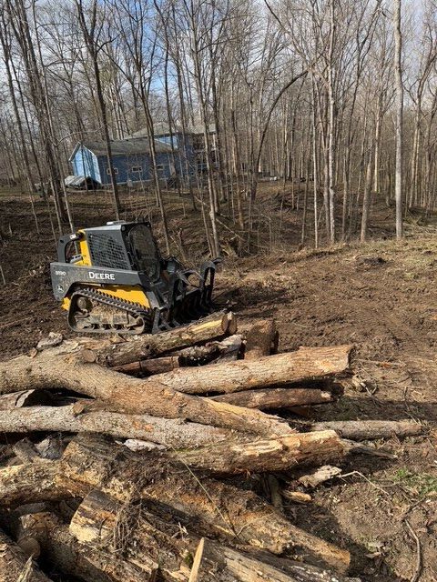 A yellow and black skid-steer loader sits in a wooded area behind a pile of felled logs, with a house visible in the rear.