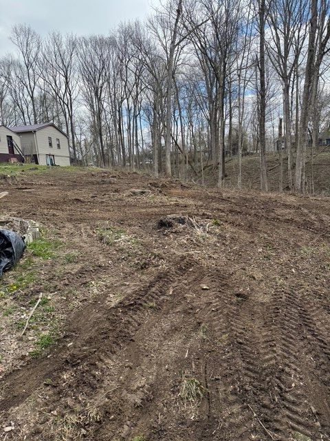 A cleared, muddy lot with tire tracks in the foreground, adjacent to a house with a forest in the background.