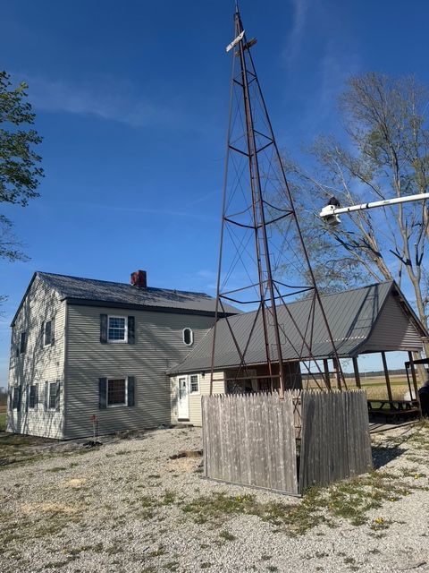 A rustic metal windmill tower stands in front of a two-story house with siding under a clear blue sky.
