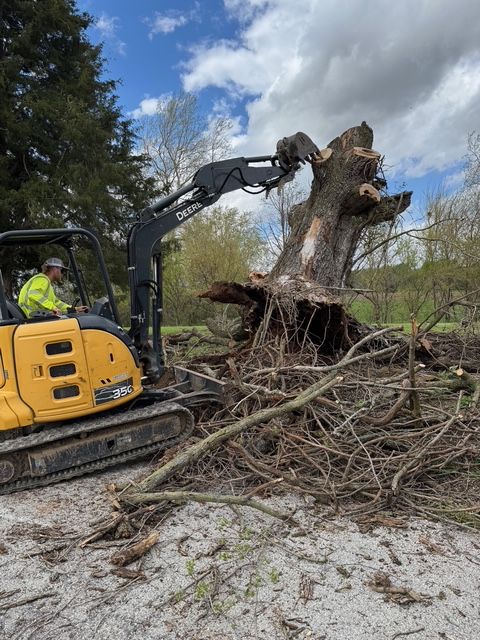 A worker operates a John Deere excavator to lift a large tree stump and root system in a rural landscape.
