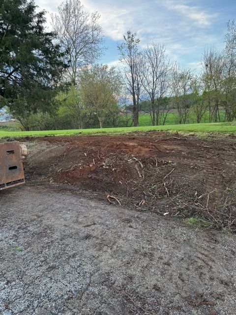 A patch of freshly cleared, churned earth next to a gravel path, with a field and trees in the background.