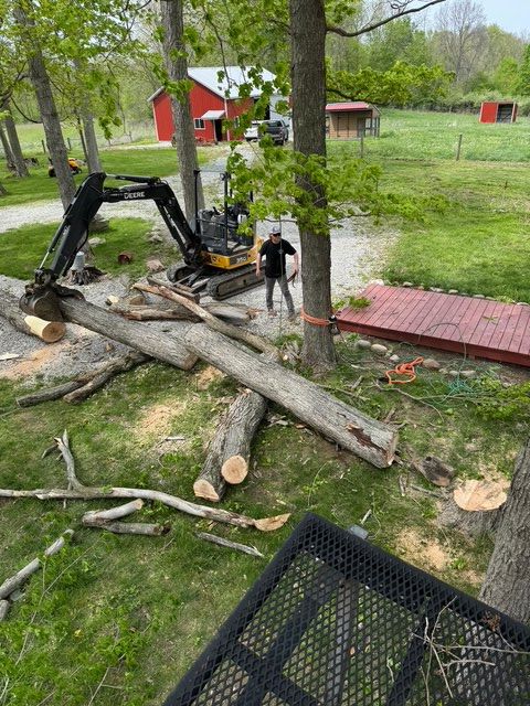 A small excavator moves a large, downed tree trunk in a grassy yard near a red barn and a deck.