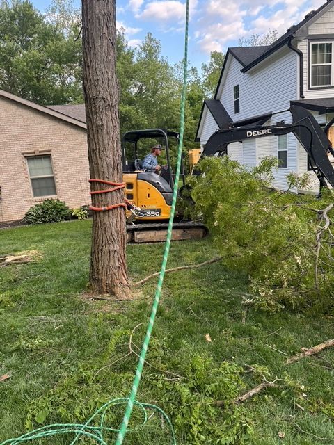 A worker operates a yellow John Deere excavator to clear fallen branches near a residential house and a tree trunk.