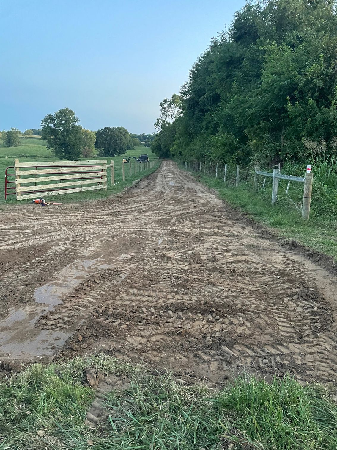 A muddy, rutted dirt path leading into a green, tree-lined landscape with wooden fencing on either side.