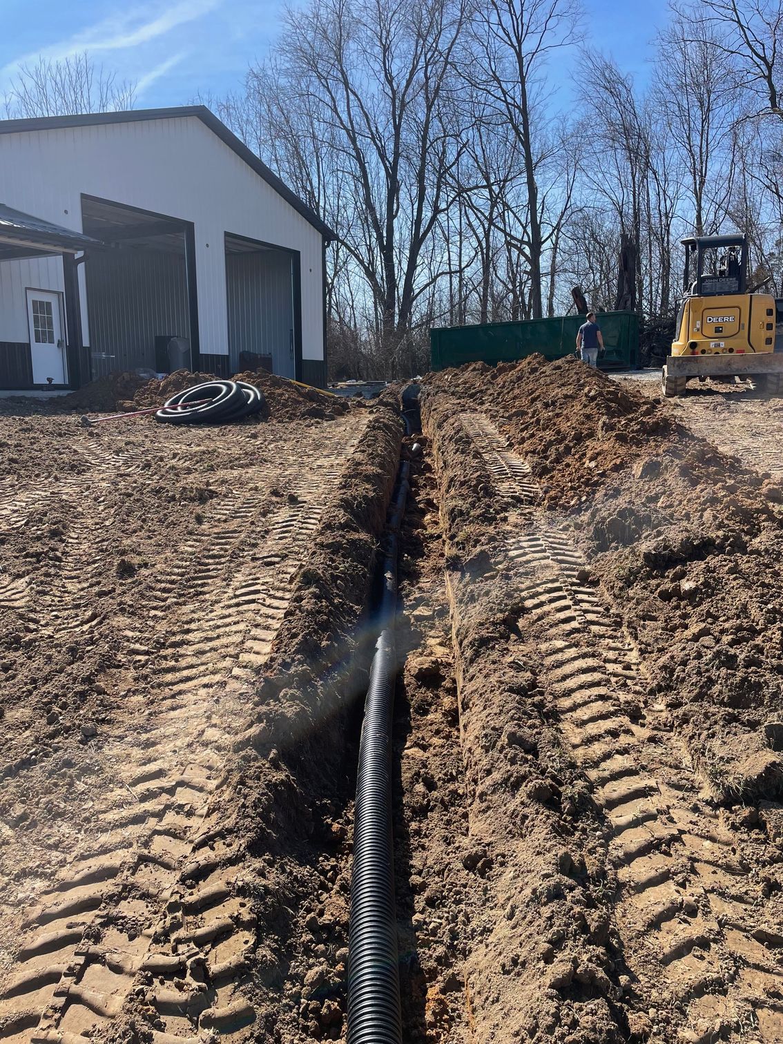 Black corrugated drainage pipe lies in a freshly dug trench leading toward a white building with a yellow excavator nearby.