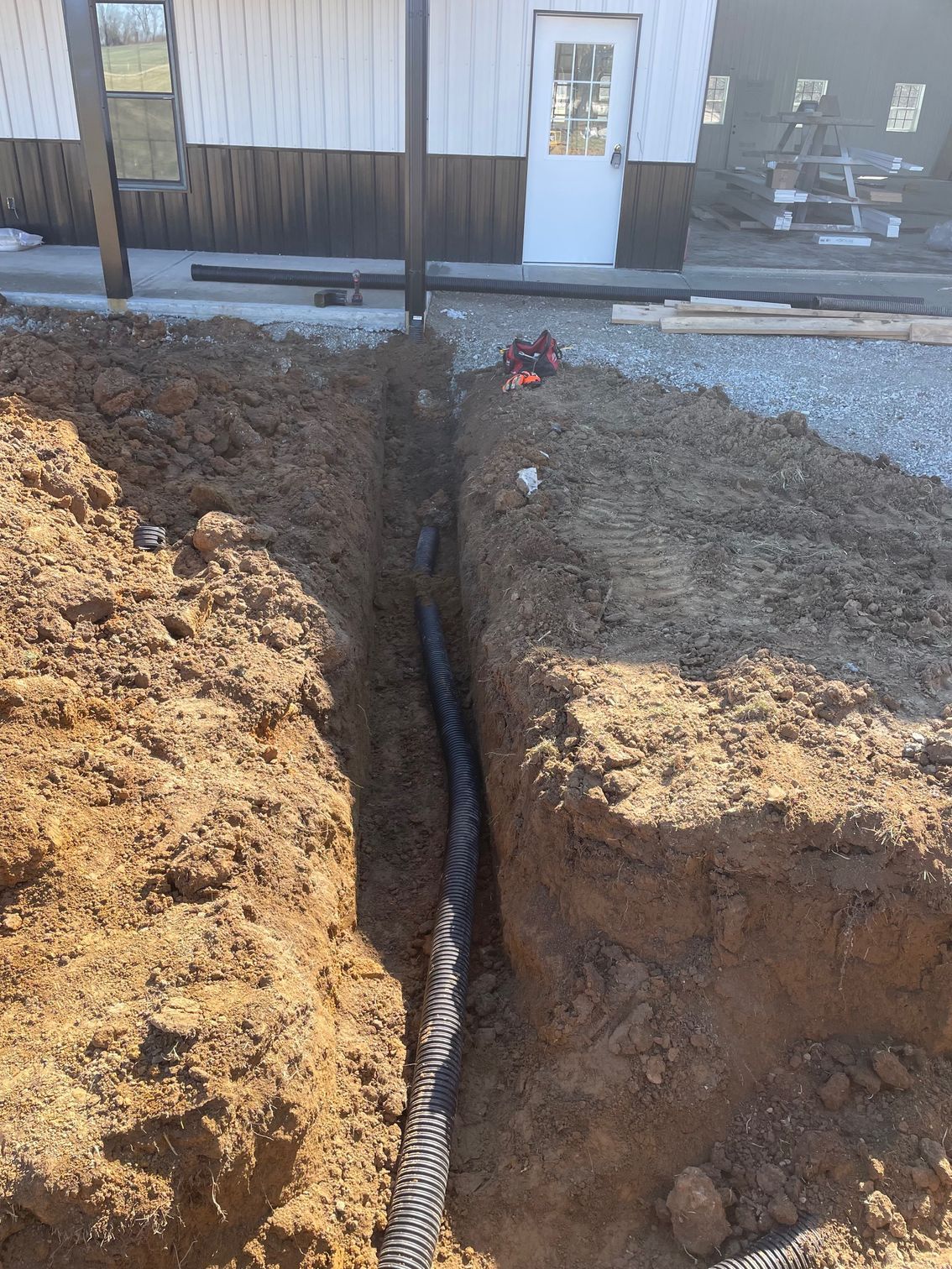 A black corrugated drainage pipe lies at the bottom of a deep, narrow dirt trench in front of a building under construction.
