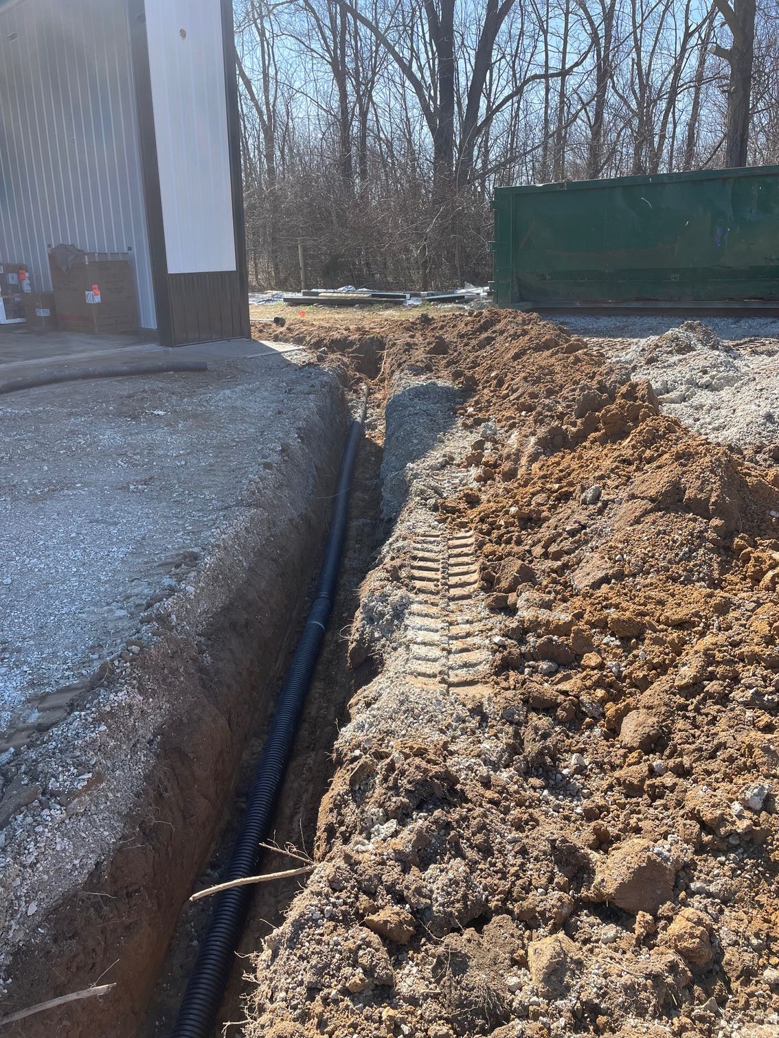 A black corrugated drainage pipe lies in a trench along the foundation of a building next to a mound of dirt.