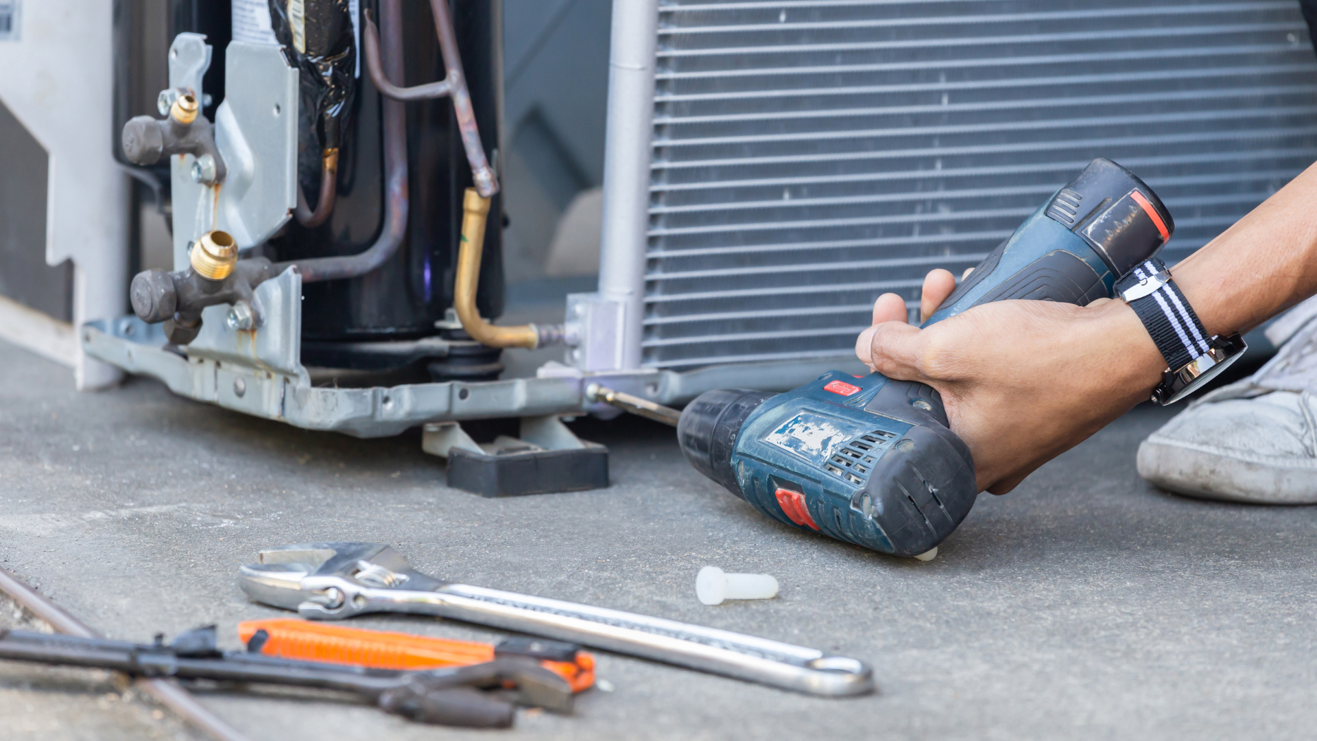 Person using a cordless drill to repair an outdoor HVAC unit, with tools on the ground.