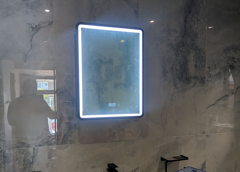 Lit mirror on a marble wall, reflection of a person reaching, inside a bathroom.