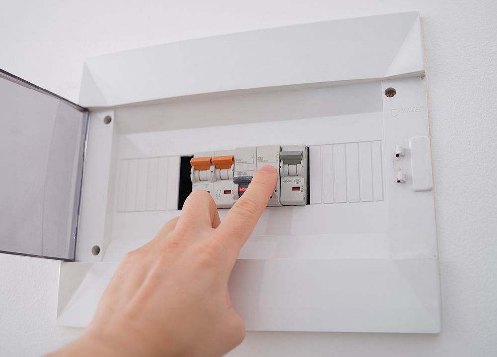 Hand flips a circuit breaker switch in a white electrical panel on a white wall.