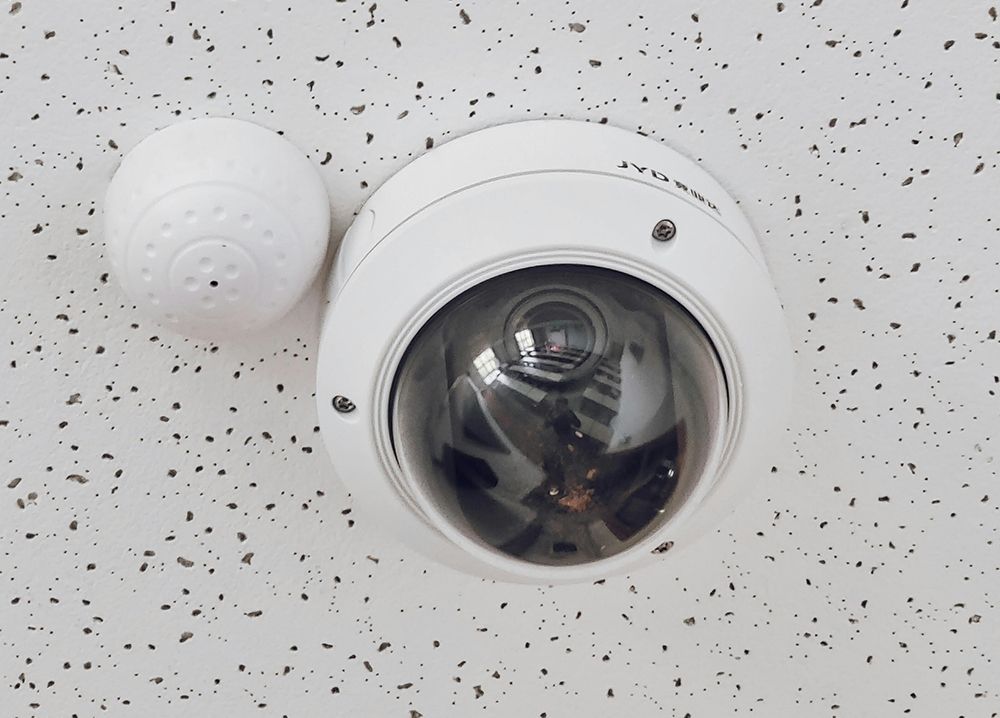 White security camera and adjacent device mounted on a speckled ceiling.